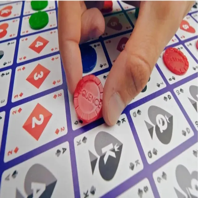 A hand placing a red chip on a card space of the Sequence board