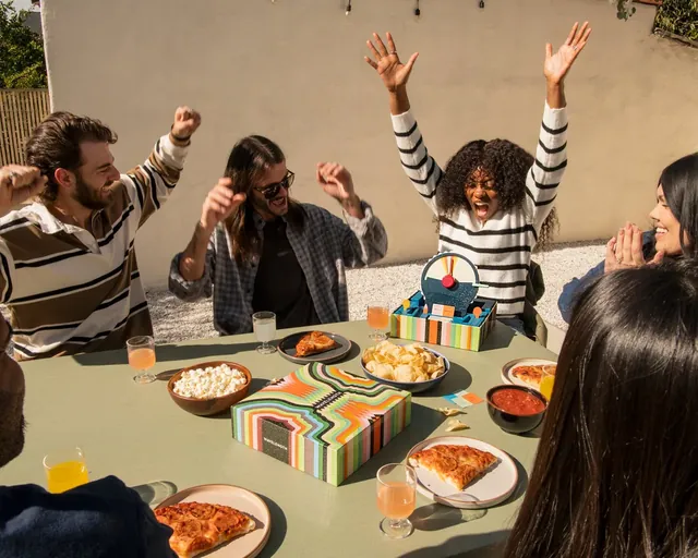 Friends at an outdoor table cheering with their arms up, the Wavelength device among plates of food