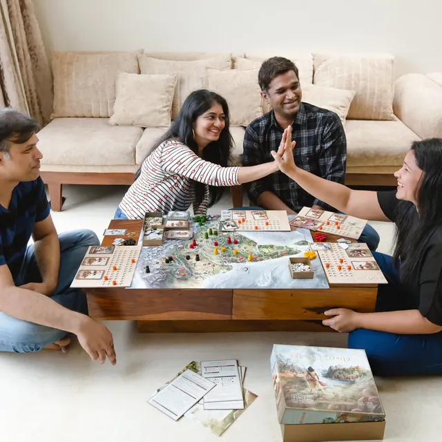 Four adults sit on the floor around a wooden coffee table with the game set up; two of them high-five across the board, and the box and rulebook lie on the floor beside them