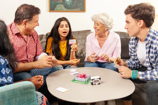 A family of five around a coffee table playing Herd Mentality, one of them reading out a question card
