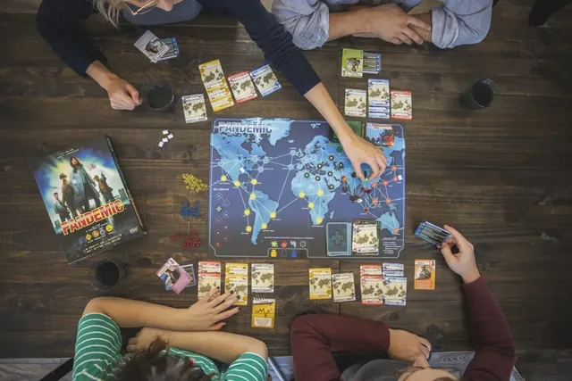 Four players around the Pandemic board, with player cards, disease cubes and pawns spread across the world map