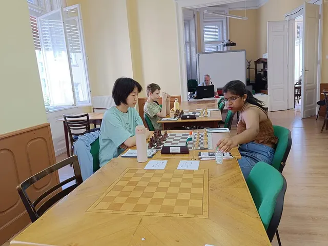 Two women playing chess at a tournament table, with other players behind them