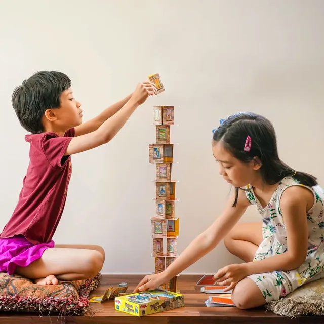 Two children sitting on the floor, taking turns to build a Rhino Hero tower