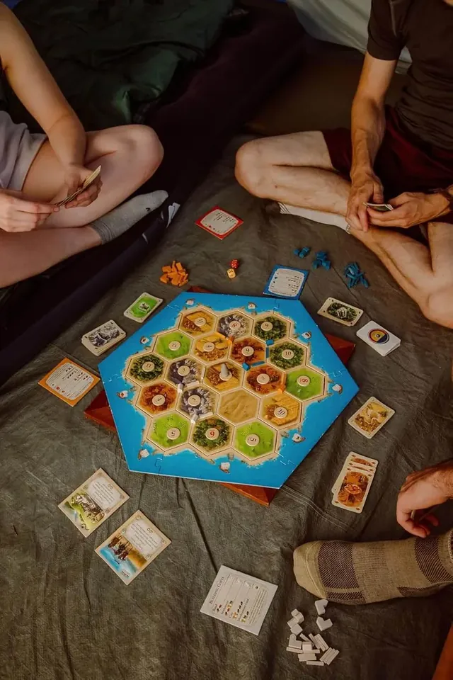Overhead view of players sitting cross-legged on a bed around the Catan board, with cards spread in front of them