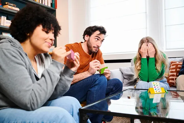 Three friends on a sofa, one covering her eyes while the others write clues
